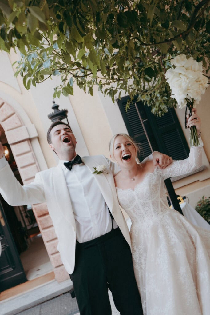 A joyful bride and groom stand outdoors, arm in arm, celebrating. The groom wears a white tuxedo jacket and the bride wears a lace wedding dress, holding a bouquet aloft. Both are smiling with mouths open in excitement.