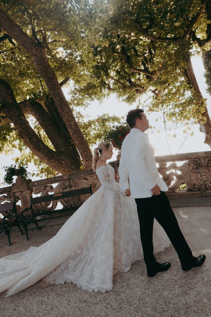 A bride in a long, lace wedding gown and a groom in a white jacket and black pants walk hand in hand outdoors under large trees, with sunlight filtering through the leaves.