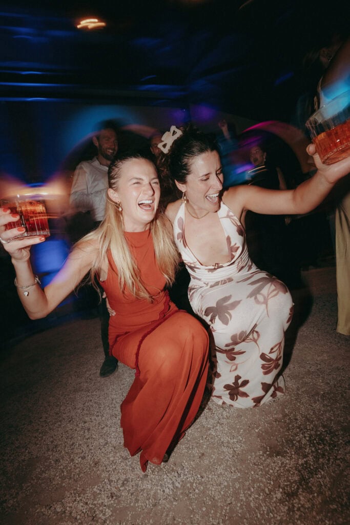 Two women kneel and laugh together at a party, holding drinks, surrounded by colorful lights and blurred people dancing in the background.