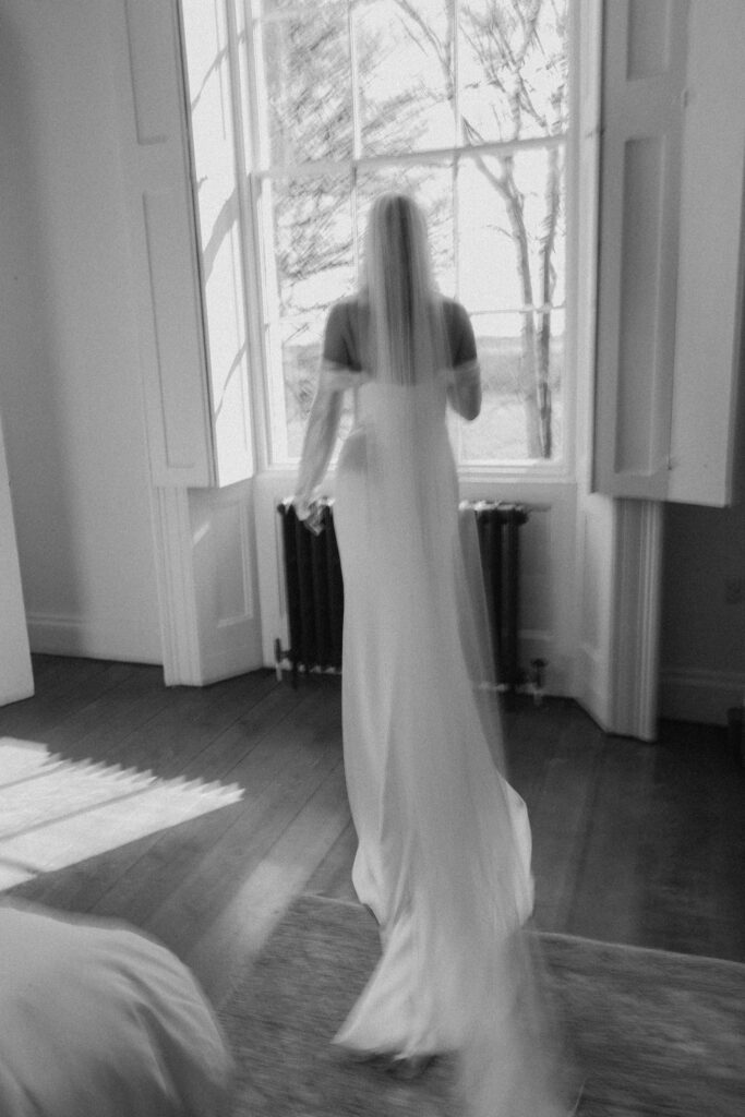 A black-and-white photo by a creative wedding photographer captures a bride in a long gown and veil near a large window, gazing outside. The scene is softly blurred for a dreamy, ethereal effect.