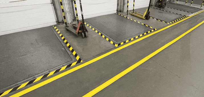 Bodenmarkierungen eines Fußwegs in RAL 1023 Verkehrsgelb, die sichere Gehwege für Mitarbeiter und Besucher gewährleisten und die Unfallgefahr reduzieren. Zusätzlich sind gelb-schwarz schraffierte Markierungen sichtbar, die vor den Rolltoren einen Gefahrenbereich kennzeichnen