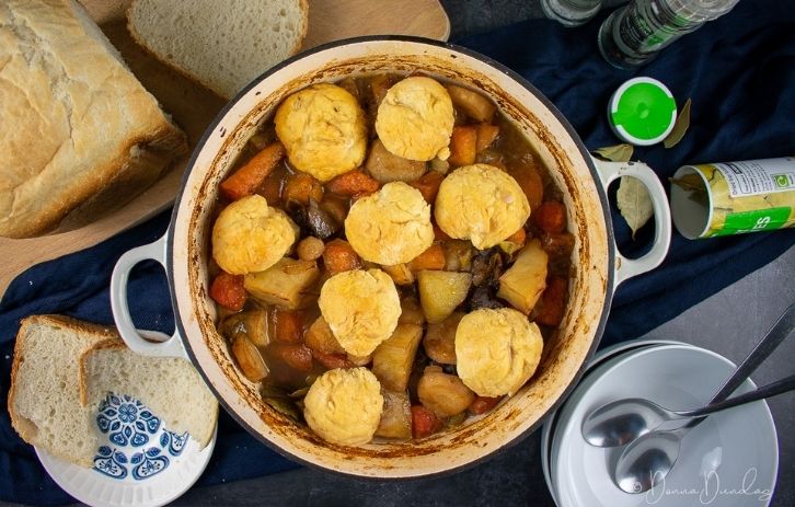 Leftover lamb stew with dumplings in a cast-iron cooking pan. Loaf of bread on chopping board above, embody bowls, bay leaves and salt and pepper to the sides.