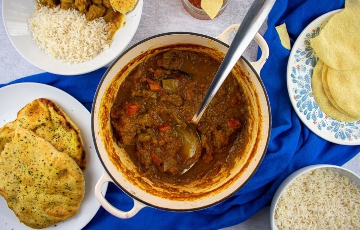 Lamb curry in cast-iron dish with serving spoon. Curry sauce brown/red, chunks of tomato and lamb visible. Plate with naan bread, poppadoms, bowl of rice and empty bowl leaving shot in the corners.