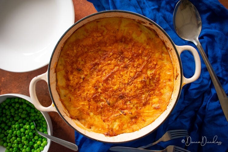 baked cottage pie in cooking dish, bowl of peas and serving spoon on blue cloth.