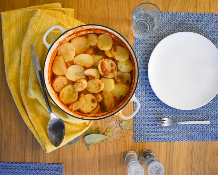 Top shot of sausage hotpot in cooking dish on table. Surrounded by plates, cutlery and salt and pepper, yellow cloth to side.