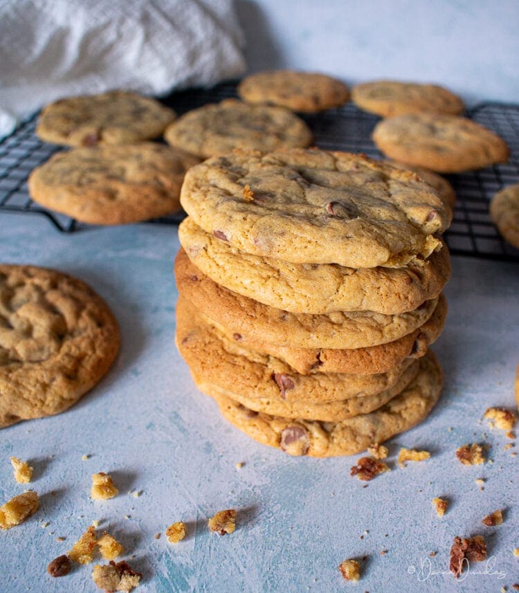 small stack of chocolate chip cookies surrounded by crumbs more cookies on wire rack in the background