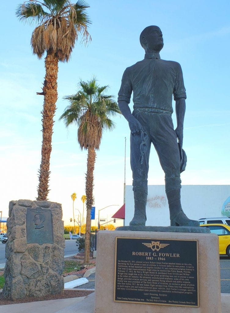 Statue of early airplane pilot holding leather googles against palm trees and cloudless sky.
