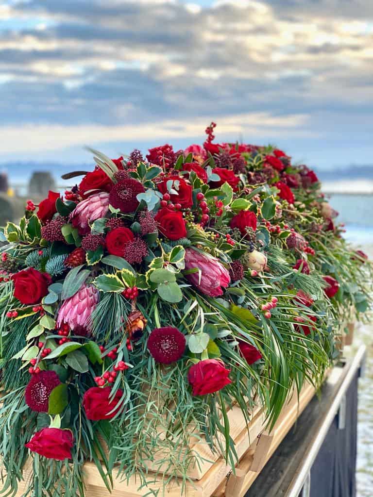 Sargbukett modern mit roten Rosen, Gerbera, Protea, Zuckerbusch, Skimmie, Ilex, Eukalyptus, Seidenkiefer auf einem Holzsarg - von Fotograf: Nicole Kemeder https://www.nickisbluetenzauber.com/