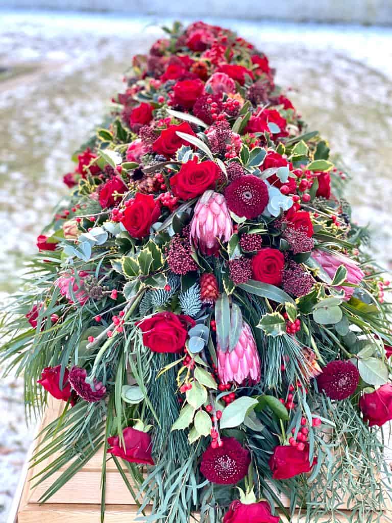 stilvolle und moderne Trauerfloristik Sargschmuck Blumenbukett für Beerdigung mit roten Rosen, Gerbera, Protea, Zuckerbusch, Skimmie, Ilex, Eukalyptus, Seidenkiefer auf einem Holzsarg - Fotograf: Nicole Kemeder https://www.nickisbluetenzauber.com/