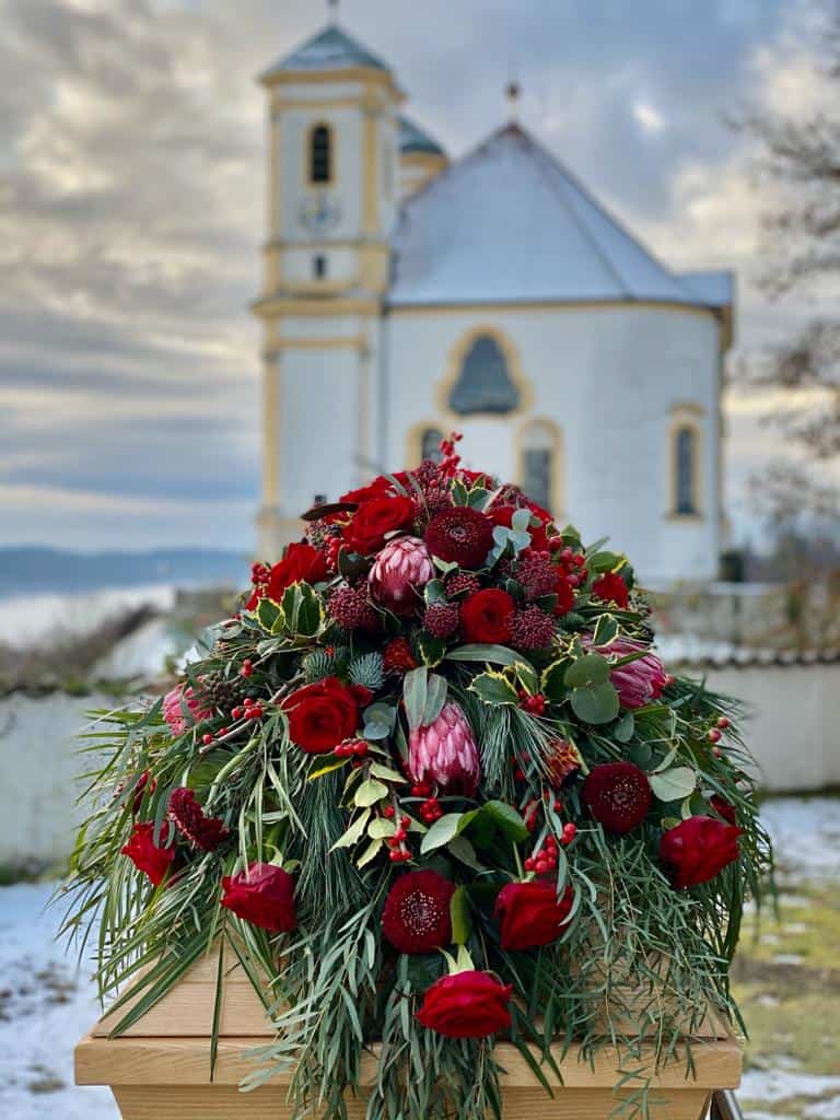 Trauerfloristik Sargblumenschmuck modern mit roten Rosen, Gerbera, Protea, Zuckerbusch, Skimmie, Ilex, Eukalyptus, Seidenkiefer auf einem Holzsarg im Hintergrund ist eine bayrische Friedhofskapelle zu sehen - Fotograf: Nicole Kemeder https://www.nickisbluetenzauber.com/