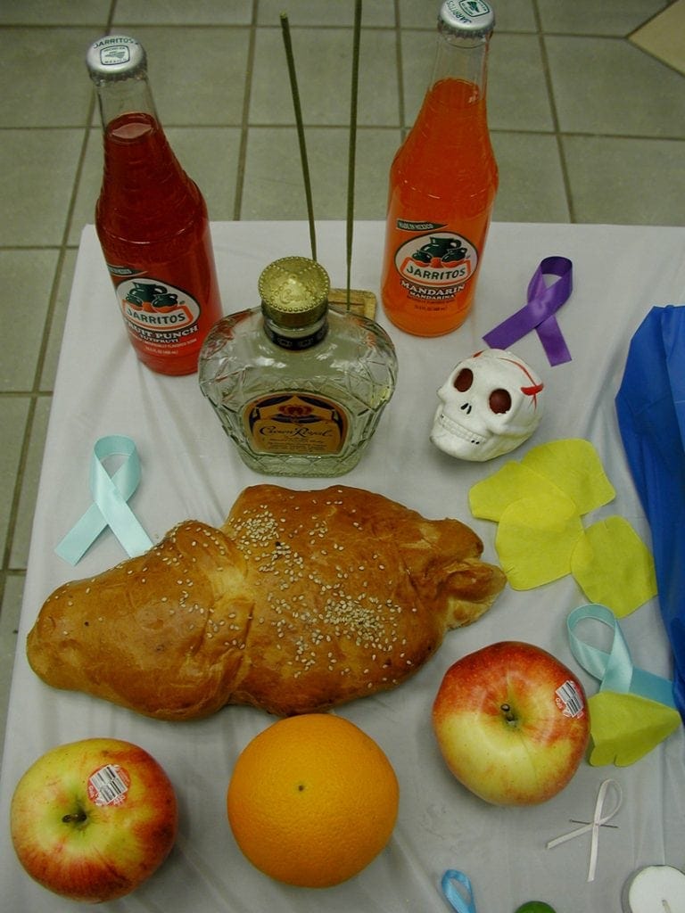Altar with food offerings including apples, Crown Royal whiskey and Day of the Dead bread