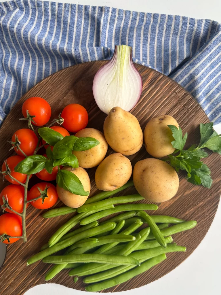 Fresh produce on a wood board with a striped towel in the back to make potato salad Italian style