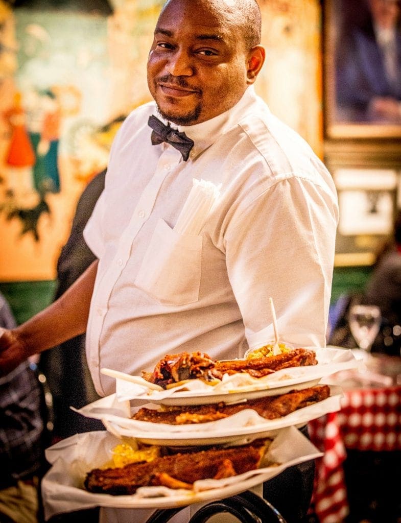 waiter in white shirt and bow tie holds 3 plate of memphis-style BBQ in one arm looking directly at camera