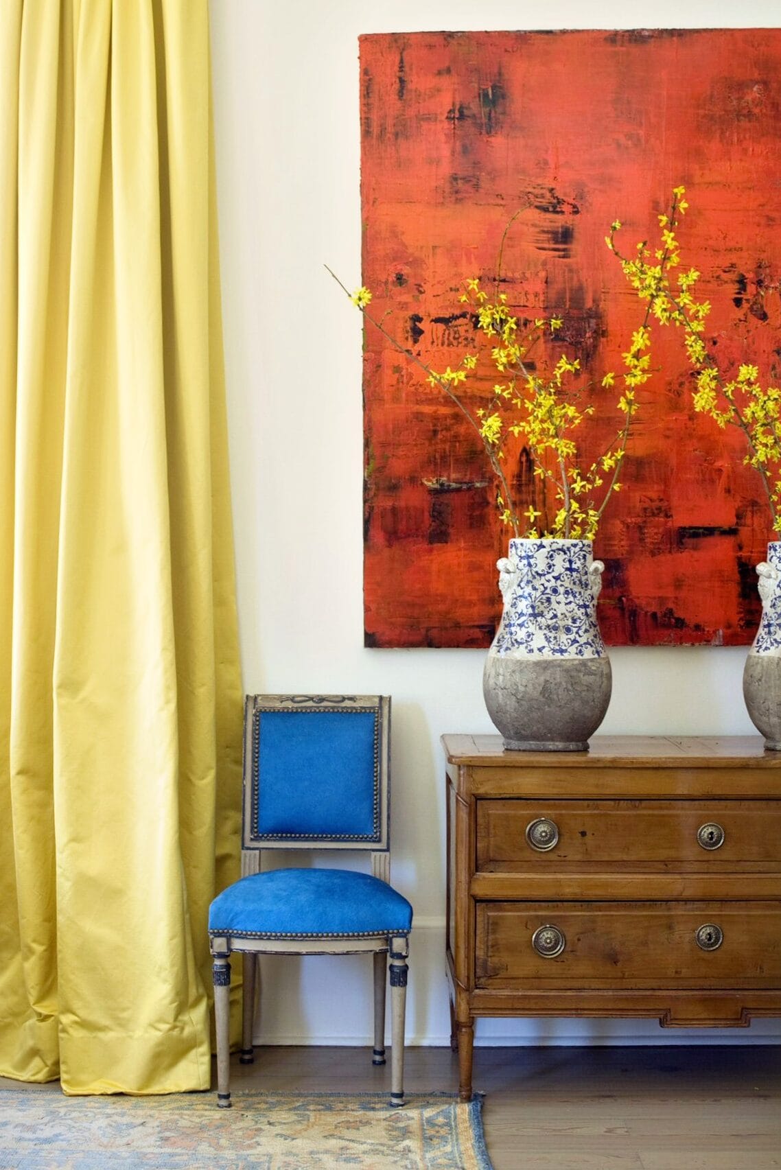 An overscaled bold red modern painting pops against Pierre Frey yellow satin curtains, cobalt blue velvet Louis XVI chairs and an antique chest. 