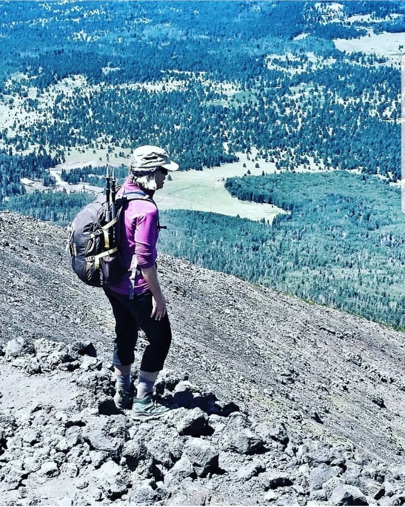 woman stands above treeline on sloping talis. Looks at forests and meadows below