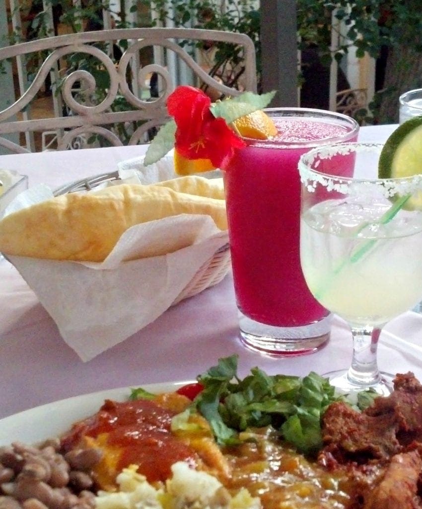 table with light pink table linens has plate of of food in foreground, margaritas and sopapilla in background - outdoors with elegant metal chair