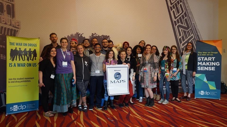 Smiling faces of SSDP members at Psychedelic Science conference. Holding a sign that says MAPS (Multidisciplinary Association for Psychedelic Studies), flanked by SSDP banners with slogans.