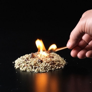 A hand holds a lit match to a pile of kindling that's now alight on a black background.