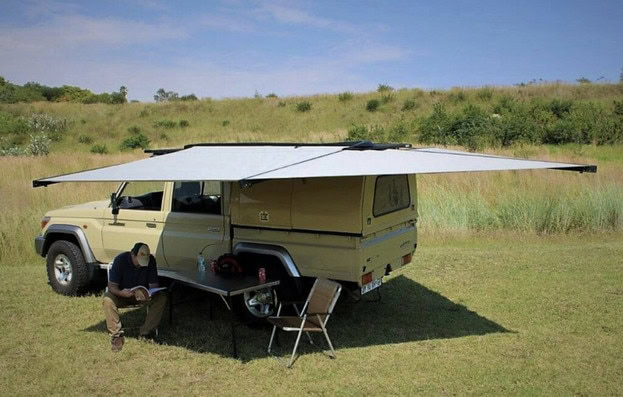 Jeep pictured in a remote field with a 270 degree awning set up and a man sat reading in the shade underneath it.