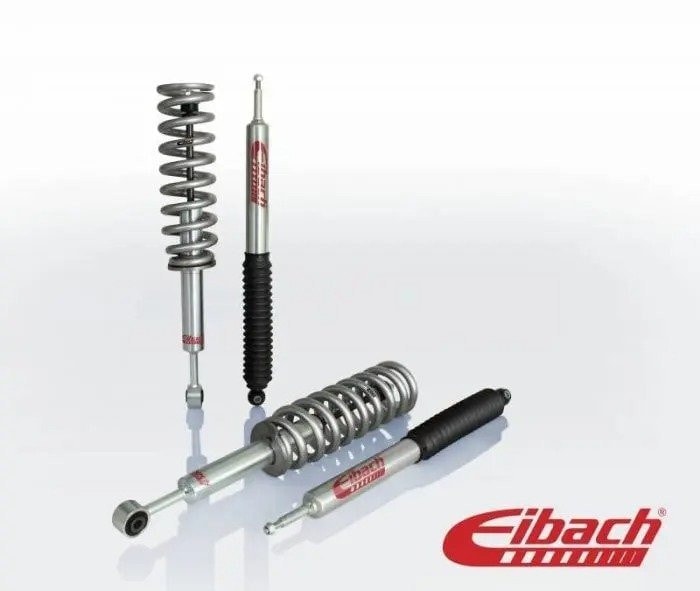 Two pairs of silver and black lift springs and shocks lie on a white surface or are balanced upright against a white background. A red Eibach logo is in the bottom right corner.