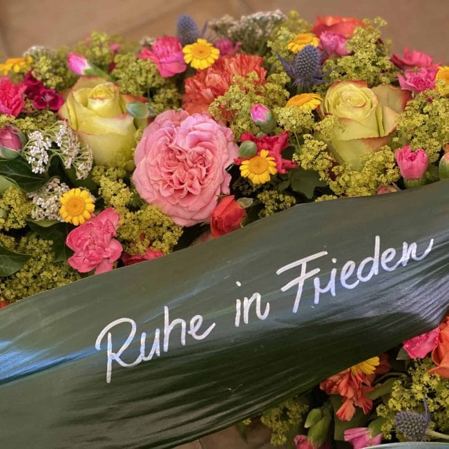 Moderner Trauerkranz ohne Schleife - mit alternative Schleife - Grünes Blatt von Schusterpalme statt Trauerschleife mit Aufschrift "Ruhe In Frieden" ca. 80 cm Durchmesser mit blauen Disteln - Mannstreu, Farbige Freiland Rosen, Frauenmantel, Schafgabe, pinke Nelken, gelbe Färberkamille, Preis ca. 380 Euro