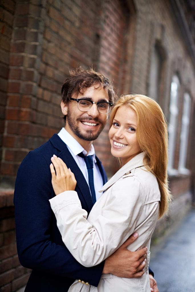 Smiling couple on a city date — man with glasses and beard, woman with reddish-brown hair, both looking at camera.