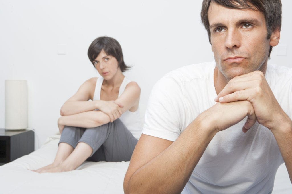 Hispanic man clasping hands under chin, looking stressed, while woman sits behind — representing breakup struggles and desire to reconnect.