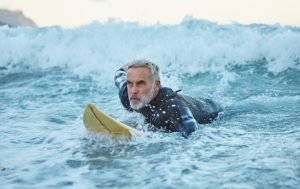 Beach, surfer and old man on surfboard swimming in nature enjoying dangerous water sports and retir