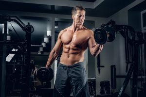 Muscular shirtless male doing biceps exercise with dumbbells.