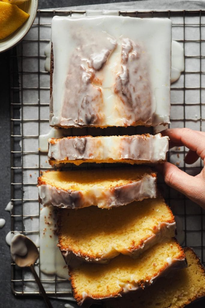 One-bowl Iced lemon ricotta loaf cake, melt-in-your-mouth tender and super moist. It's easy to make and the perfect treat! A hand is picking up a slice | Paula's Apron Recipe