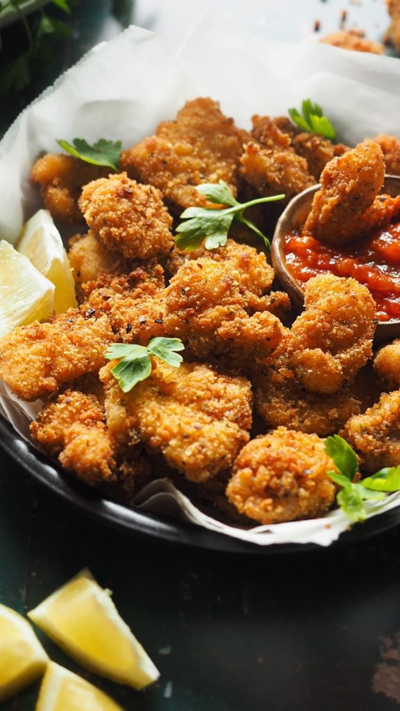 Nuggets de Pescado en 15 Minutos, crujientes, fáciles y que enamoran a todos los públicos. Con salsa de tomate casera son la perdición. En esta foto a 45 grados se ven los nuggets de rape con un rebozado dorado crujiente servidos en un plato con perejil por encima, gajos de limón alrededor y un bol pequeño con salsa de tomate para mojar. Dentro del bol hay un nugget de rape. | Receta de Paula's Apron