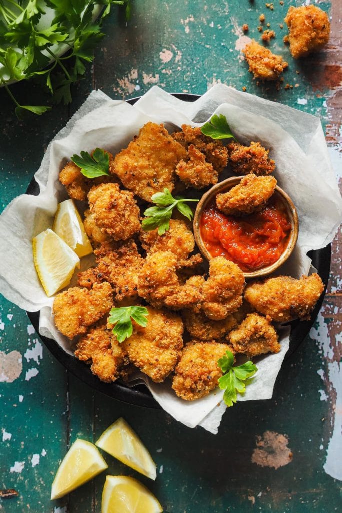 Nuggets de Pescado en 15 Minutos, crujientes, fáciles y que enamoran a todos los públicos. Con salsa de tomate casera son la perdición. En esta foto cenital se ven los nuggets de rape con un rebozado dorado crujiente servidos en un plato con perejil por encima, gajos de limón alrededor y un bol pequeño con salsa de tomate para mojar. Dentro del bol hay un nugget de rape. | Receta de Paula's Apron