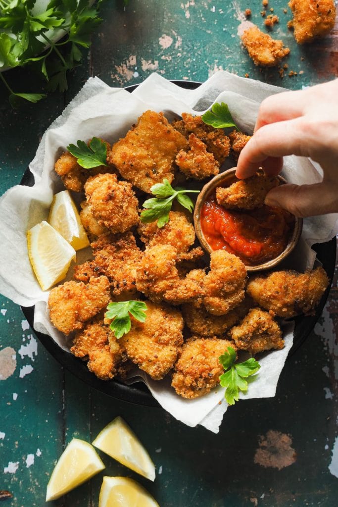 Nuggets de Pescado en 15 Minutos, crujientes, fáciles y que enamoran a todos los públicos. Con salsa de tomate casera son la perdición. En esta foto cenital se ven los nuggets de rape con un rebozado dorado crujiente servidos en un plato con perejil por encima, gajos de limón alrededor y un bol pequeño con salsa de tomate para mojar. Una mano está mojando un nugget de pescado en la salsa. | Receta de Paula's Apron