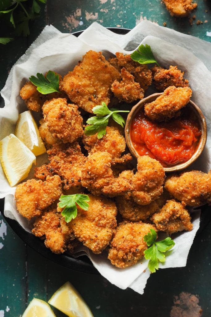 Nuggets de Pescado en 15 Minutos, crujientes, fáciles y que enamoran a todos los públicos. Con salsa de tomate casera son la perdición. En esta foto cenital se ven los nuggets de rape con un rebozado dorado crujiente servidos en un plato con perejil por encima, gajos de limón alrededor y un bol pequeño con salsa de tomate para mojar. Dentro del bol hay un nugget de rape. | Receta de Paula's Apron