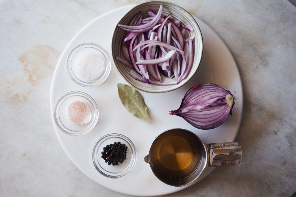 Hacer Cebolla Encurtida casera no puede ser más fácil. Es otro básico de cocina que no puede faltar en nuestro repertorio por su versatilidad y es que si encima usamos cebolla morada el color que queda es otro nivel. ¿Consecuencia? No solo aportará un toque extra a tus platos, sino que además les dará un toque de color bestial. Se requieren muy poquitos ingredientes (cebolla, vinagre, pimienta, laurel, sal y azúcar en la foto), un poco de paciencia para el reposo y como resultado un encurtido increíble. | Receta de Paula's Apron