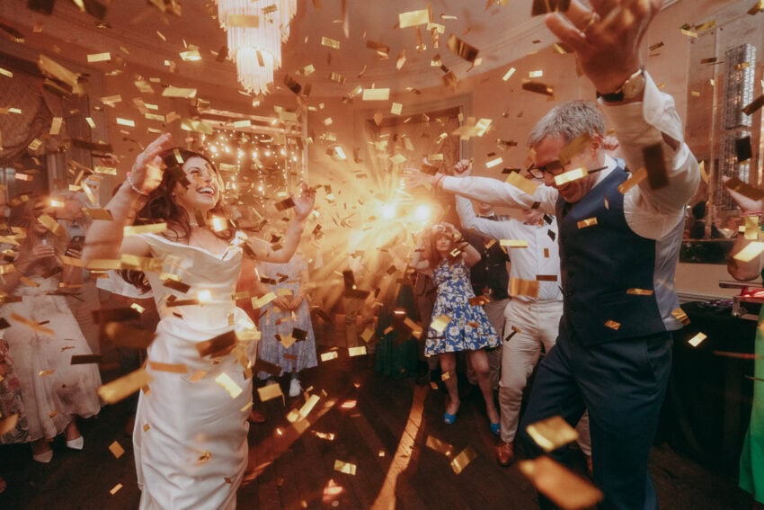 A bride and groom dance joyfully among guests as golden confetti falls around them, with warm light shining in the background—captured perfectly by a wedding photographer at a celebratory indoor wedding reception.
