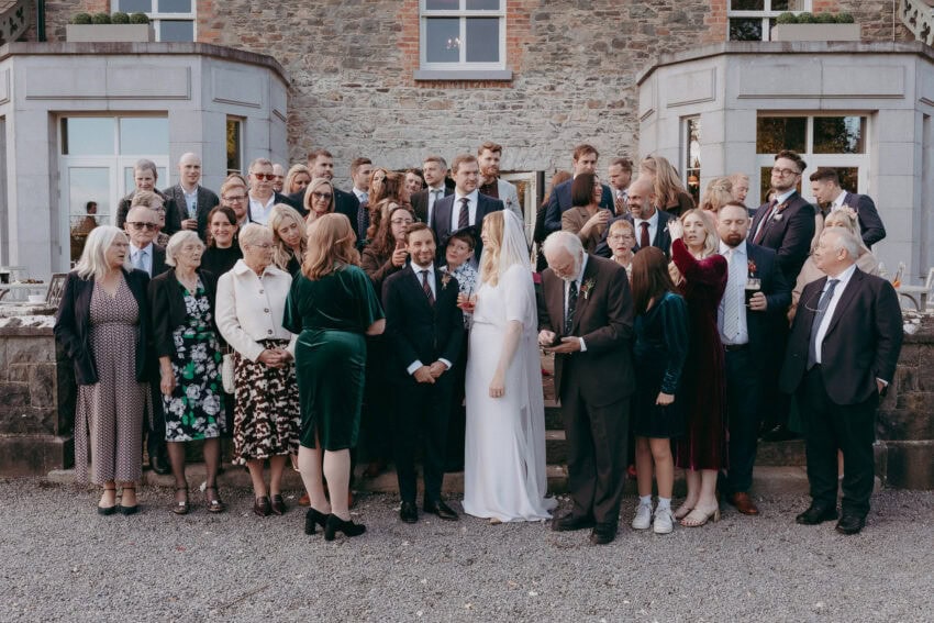 A large group of people dressed in formal attire pose for a photo outside a stone building, with the bride and groom in the center, captured perfectly by a Creative Wedding Photographer surrounded by family and friends.