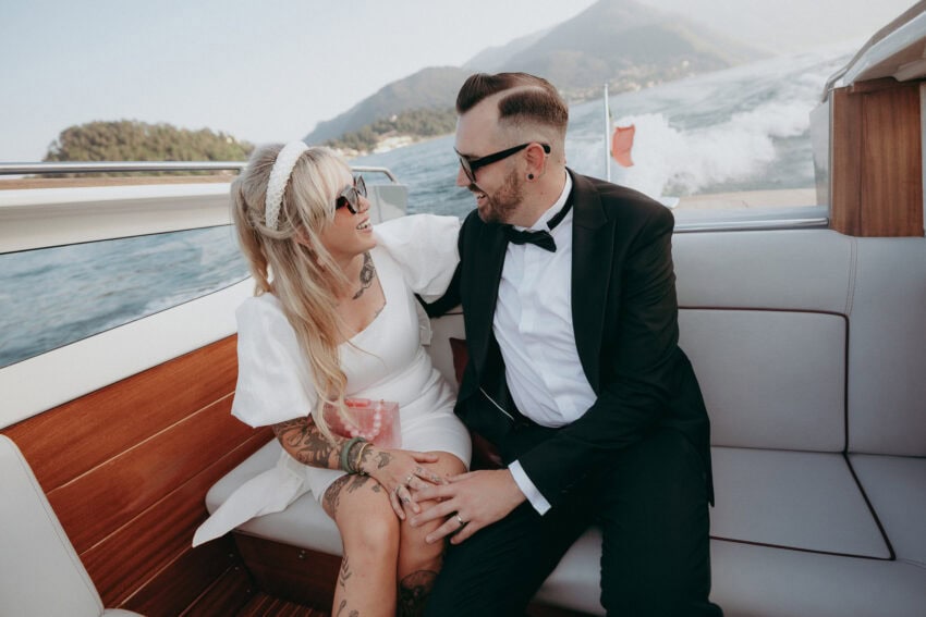 A couple dressed in formal attire, with the woman in a white dress and the man in a tuxedo, sit close together smiling on a boat as a wedding photographer captures the moment, with mountains and water in the background.