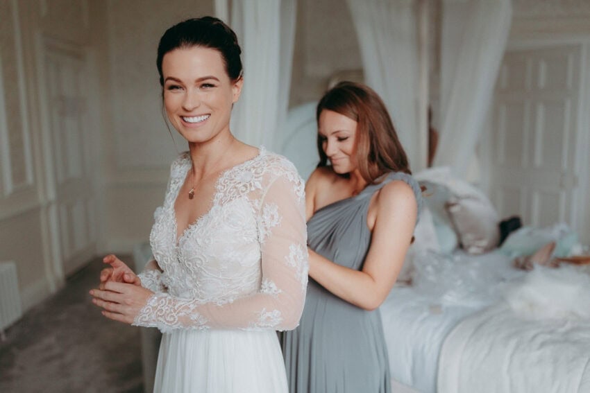 A smiling bride in a lace wedding dress stands as her Creative Wedding Photographer captures the moment a bridesmaid in a gray dress helps fasten her gown in a softly lit, elegant bedroom.