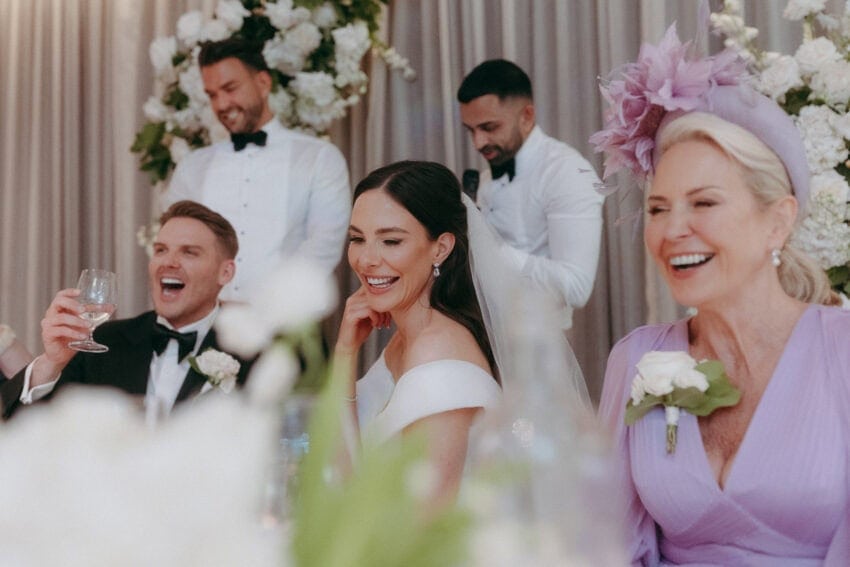 A bride and groom laugh joyfully at a wedding reception, captured by a creative wedding photographer. They sit beside a smiling woman in lavender as two men speak behind them, with white flowers and drapes decorating the background.