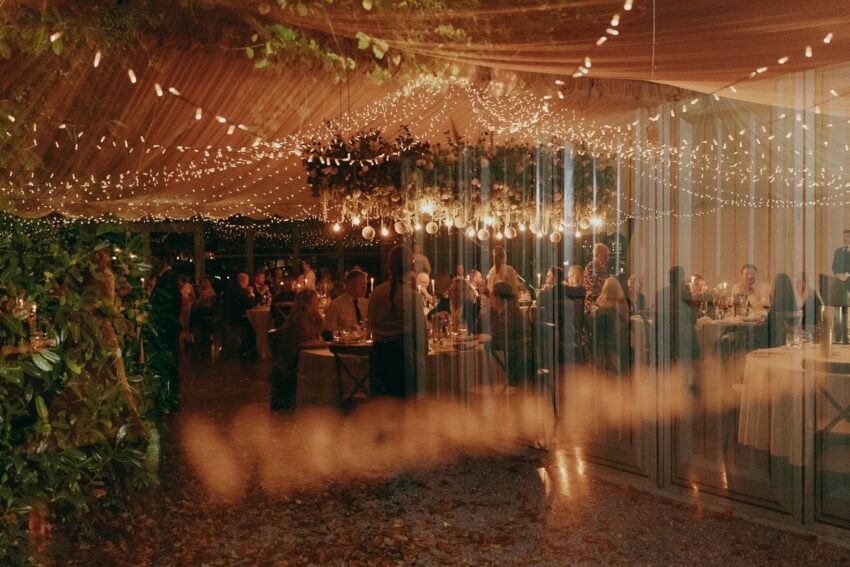 A warmly lit indoor wedding reception captured by a creative wedding photographer, with guests seated at round tables beneath strings of fairy lights and hanging greenery. Reflections shimmer on a glass surface in the festive, romantic atmosphere.