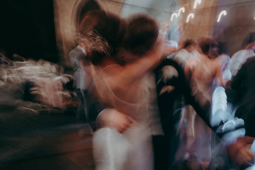A blurry, motion-filled photo by a creative wedding photographer captures two people embracing and dancing in formal attire, surrounded by other figures in a dimly lit indoor setting, evoking a joyful, energetic atmosphere.