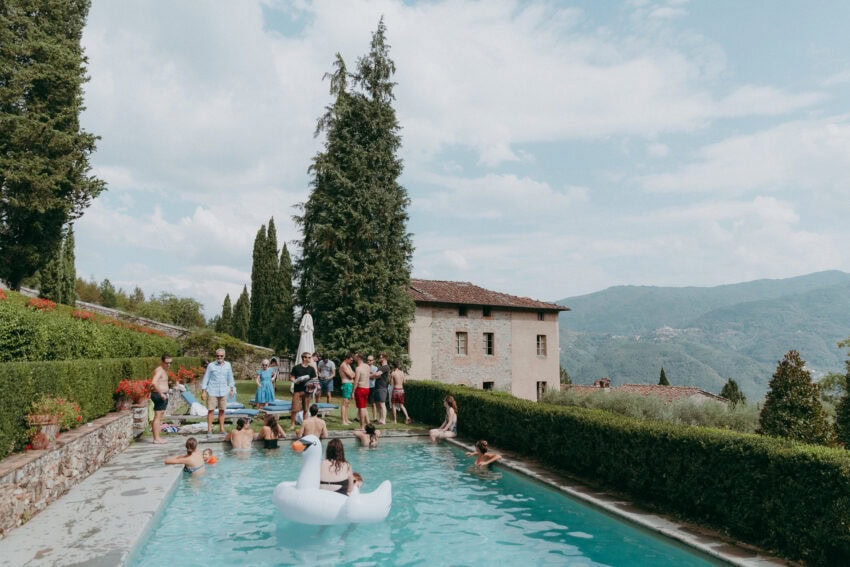 A group of people gather around and swim in a rectangular outdoor pool with a large swan float. Perfect for a Creative Wedding Photographer, the scene features tall trees, hedges, a rustic stone house, rolling hills, and a blue sky.