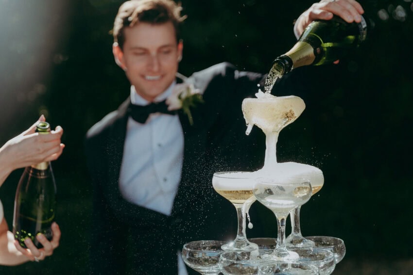A Creative Wedding Photographer captures a person in a tuxedo pouring champagne into a tower of coupe glasses, with sunlight highlighting the joyous, bubbly overflow and the celebratory atmosphere.