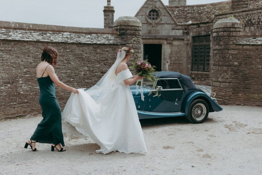 A Creative Wedding Photographer captures a bride in a white dress and veil, holding a bouquet as a woman in a dark green dress helps with her train. They walk toward a vintage blue car parked before a charming stone building.