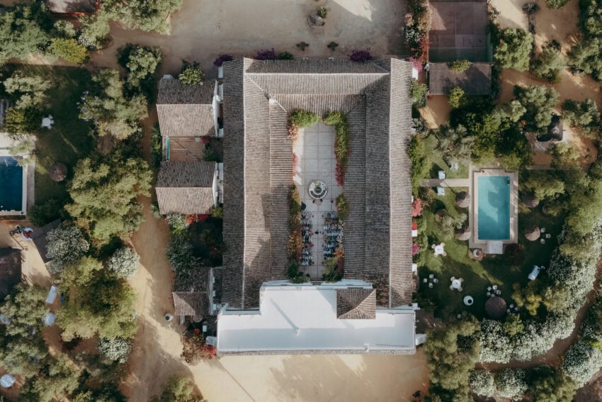 Aerial view of a rectangular villa with a tiled roof, central courtyard and fountain, surrounded by greenery. Perfect for a wedding photographer, the gardens feature a swimming pool and scattered outdoor seating around the building.