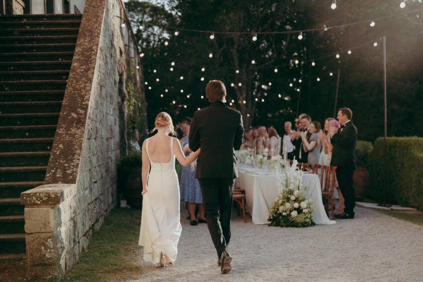 A bride and groom walk hand in hand toward an outdoor reception, where guests stand and applaud near a long, elegantly decorated table under string lights—a perfect scene for a Creative Wedding Photographer to capture in a garden setting.