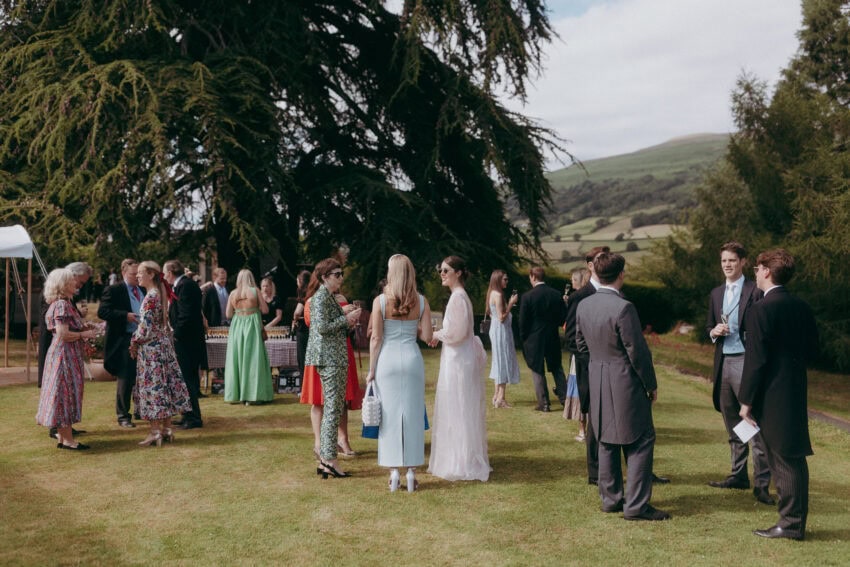 A group of well-dressed people gather and chat outdoors on a lawn with trees and hills in the background, while a wedding photographer captures candid moments at this elegant formal event or wedding reception.