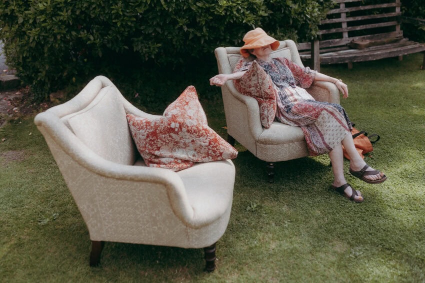 An older woman in a patterned dress and orange hat relaxes on an armchair outdoors, her arm resting nearby. Captured by a wedding photographer, the empty floral-pillow chair beside her adds charm to the serene grassy lawn setting.