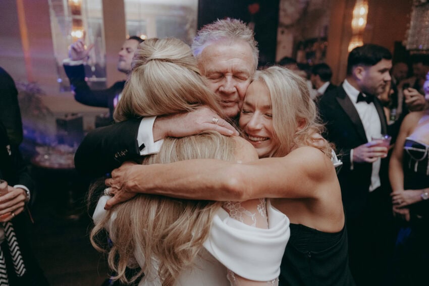Three people, dressed formally, share a joyful group hug and smile at a lively indoor event. A wedding photographer captures the moment as other guests in suits and elegant attire celebrate in the warmly lit background.